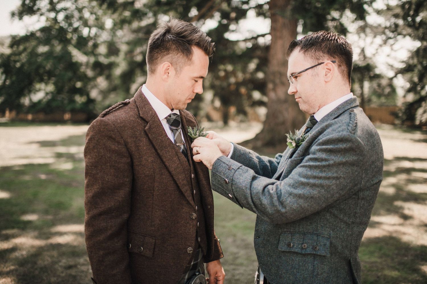 Two men in traditional Scottish formal wear adjusting each other's attire outdoors.