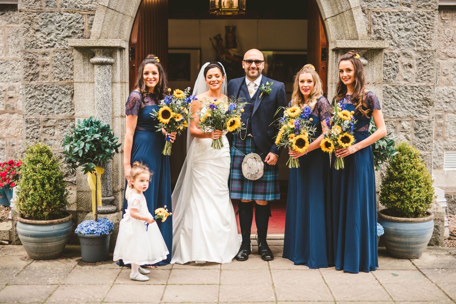 Wedding party with sunflower bouquets poses outside stone church entrance.