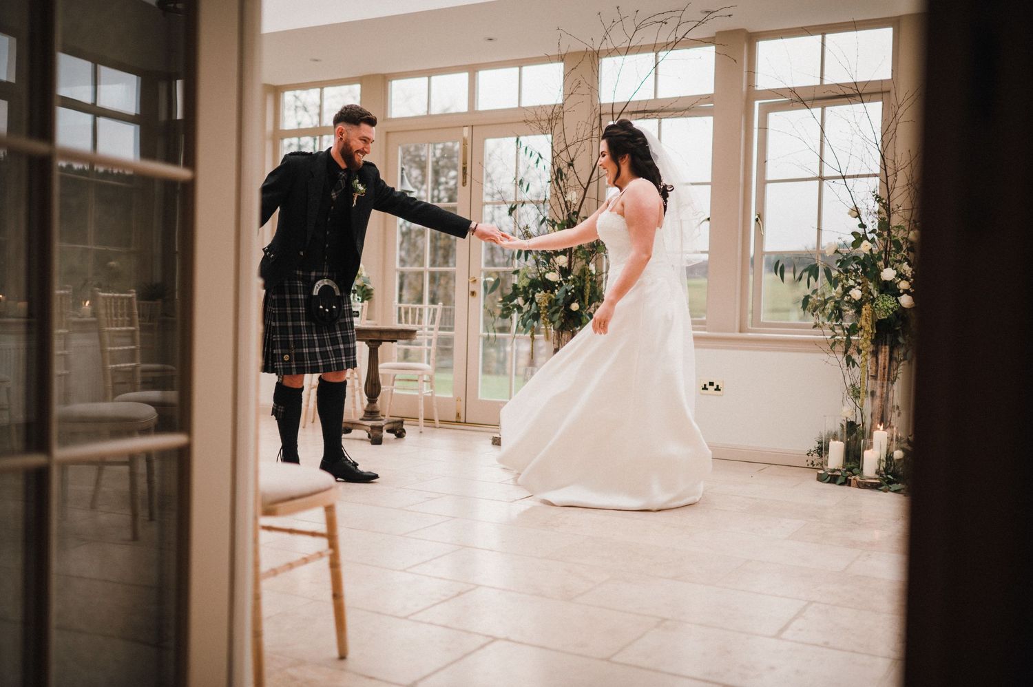 Couple performs playful dance moves in sunlit wedding venue with floor-length windows.