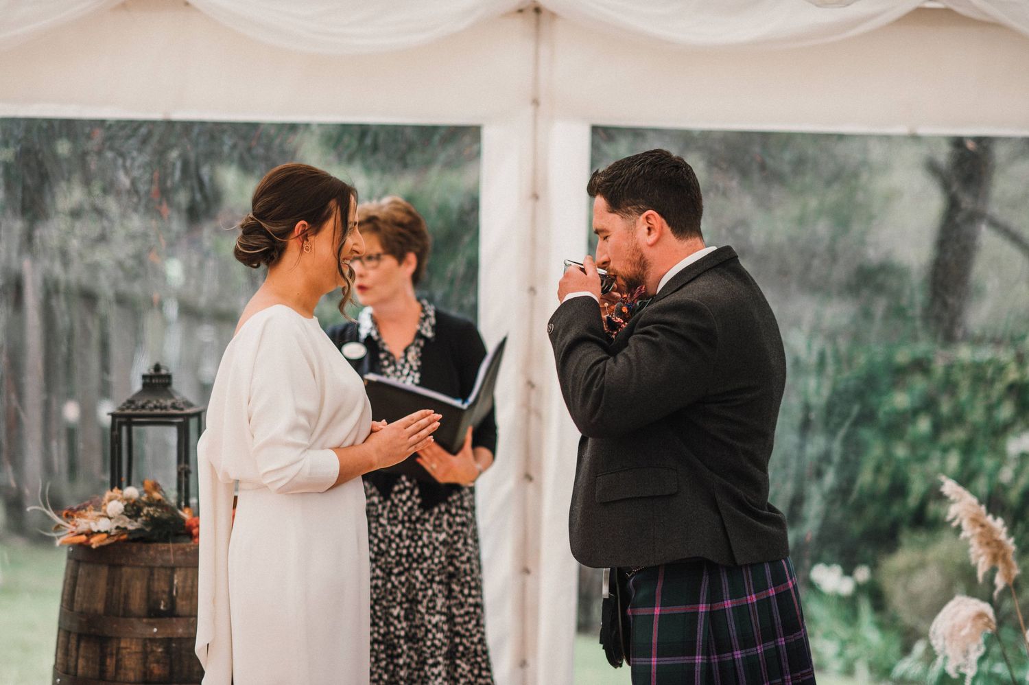 Wedding ceremony in progress under white tent with rustic barrel and lantern decorations.