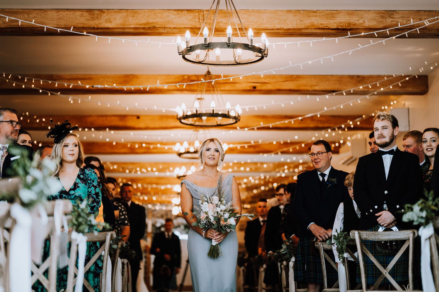 Wedding ceremony in a rustic wooden barn venue with hanging lights and white cross-back chairs lining the aisle.