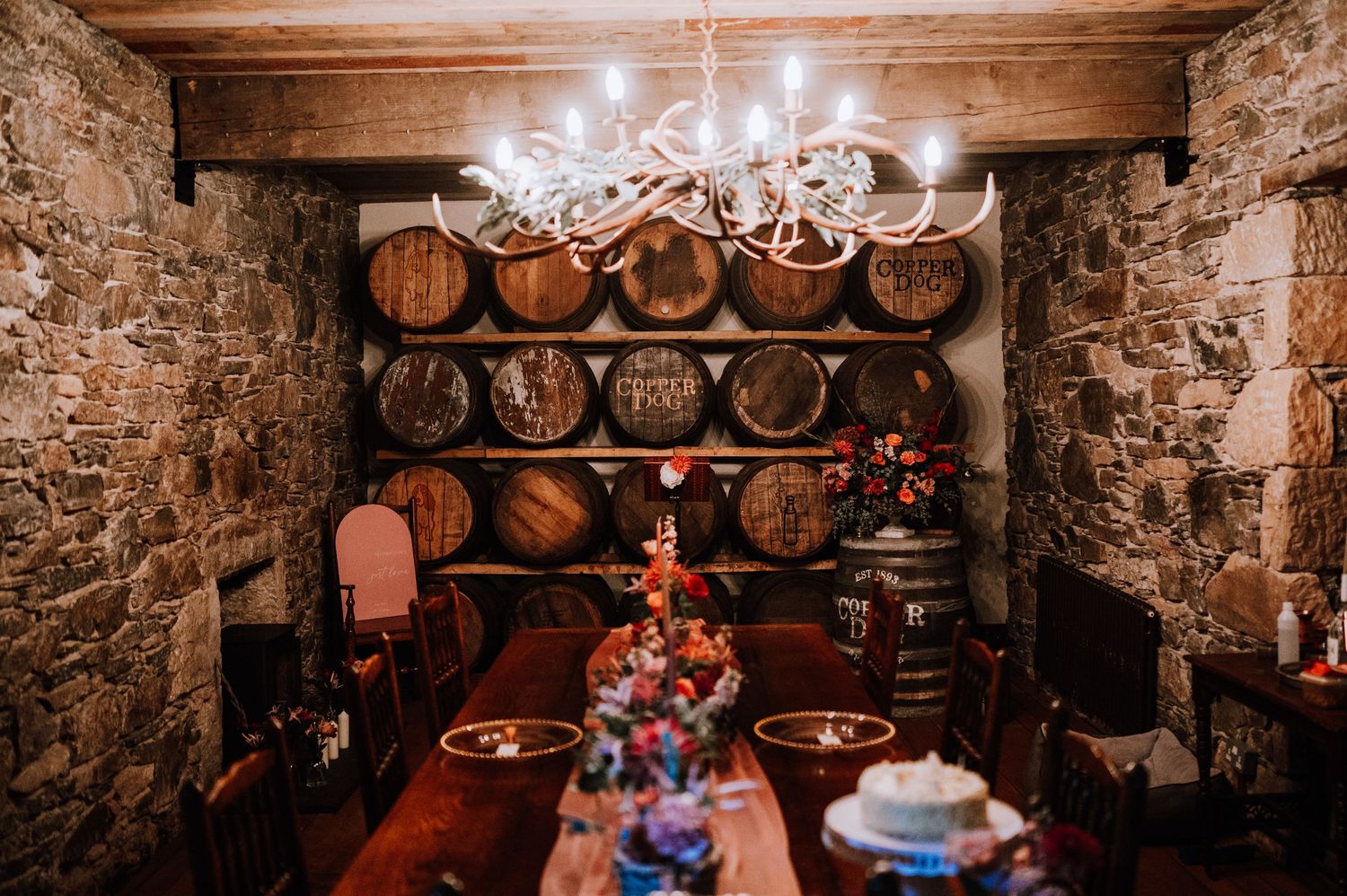 Wine cellar wedding reception setup with chandelier lighting and wooden barrel wall backdrop.