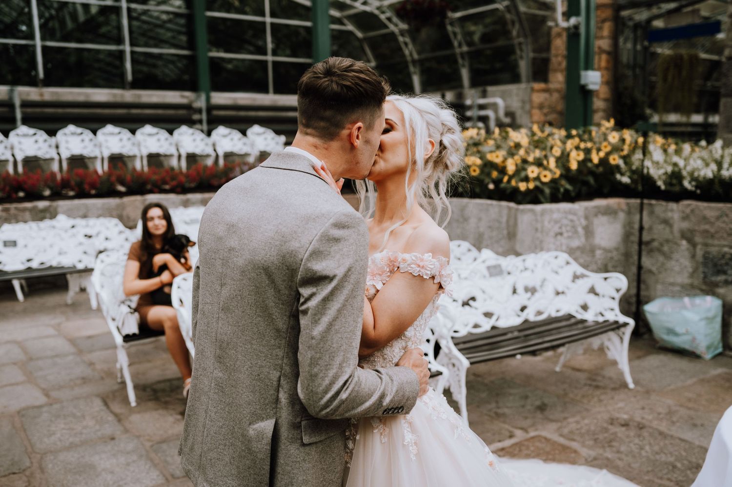 A romantic wedding moment captured in a greenhouse setting with white benches and greenery.