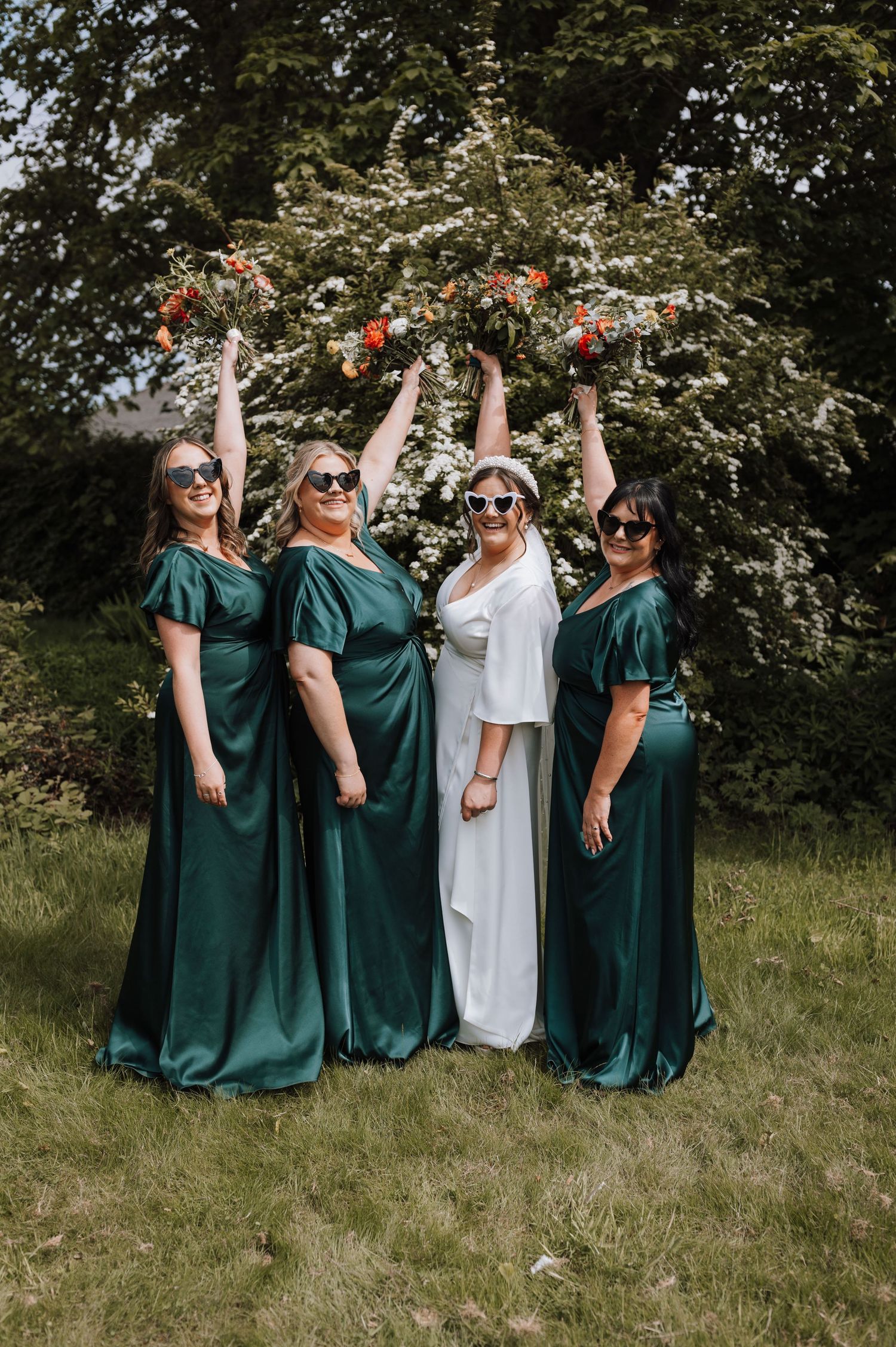 Bridal party in emerald dresses holding bouquets up in a playful outdoor garden pose.