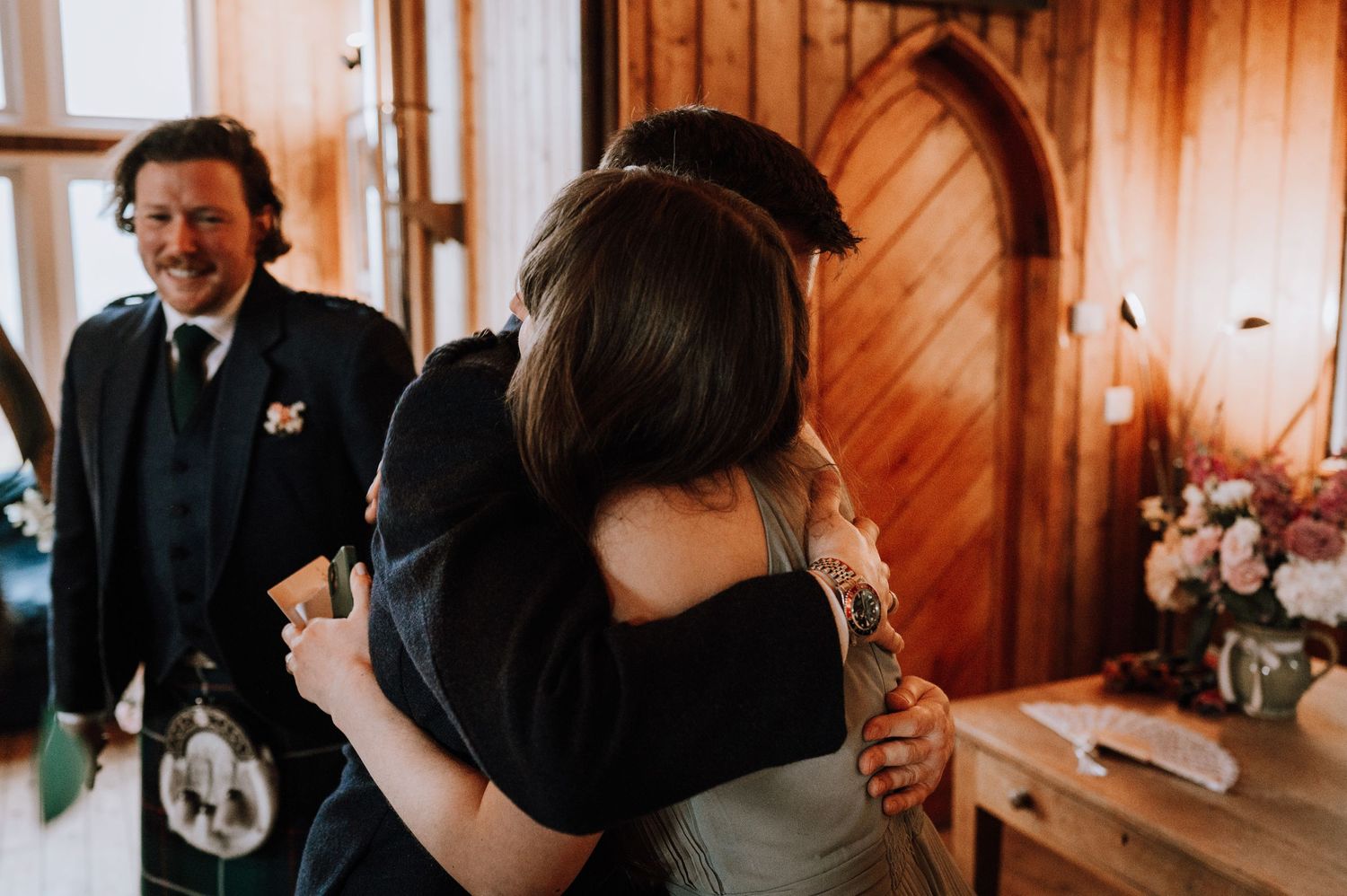 Wedding guests share emotional hugs in a cozy wood-paneled room decorated with floral arrangements.