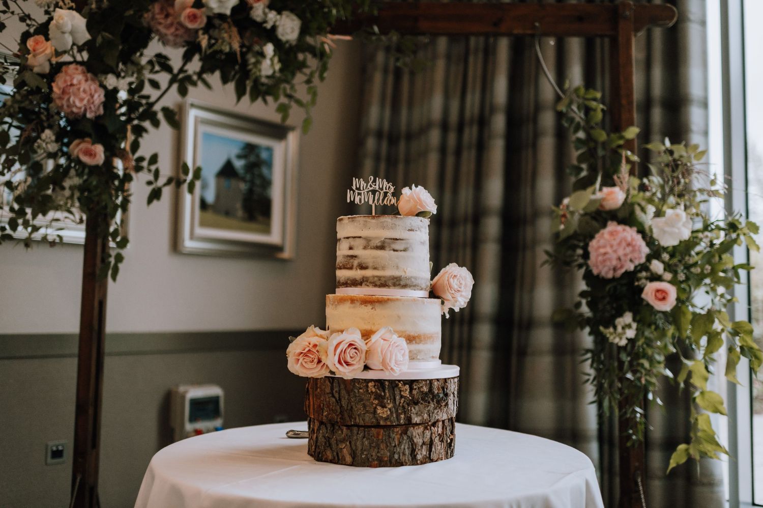 A rustic wedding cake displayed on a wooden log slice, decorated with blush flowers and surrounded by floral arrangements.