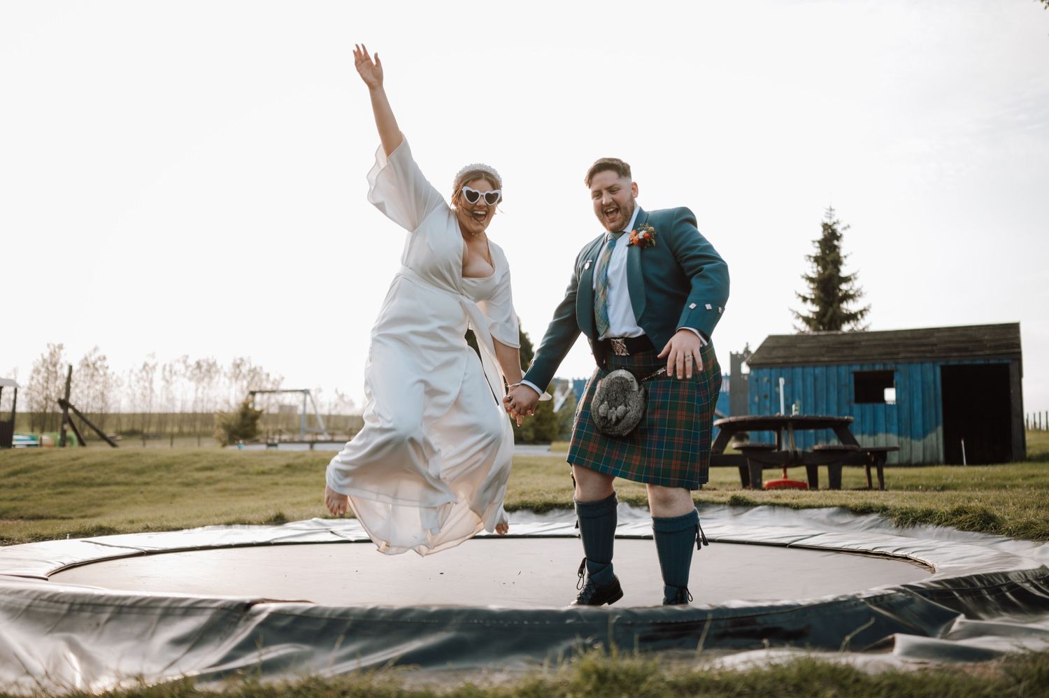 Newlyweds bounce joyfully on outdoor trampoline with groom in traditional Scottish kilt.