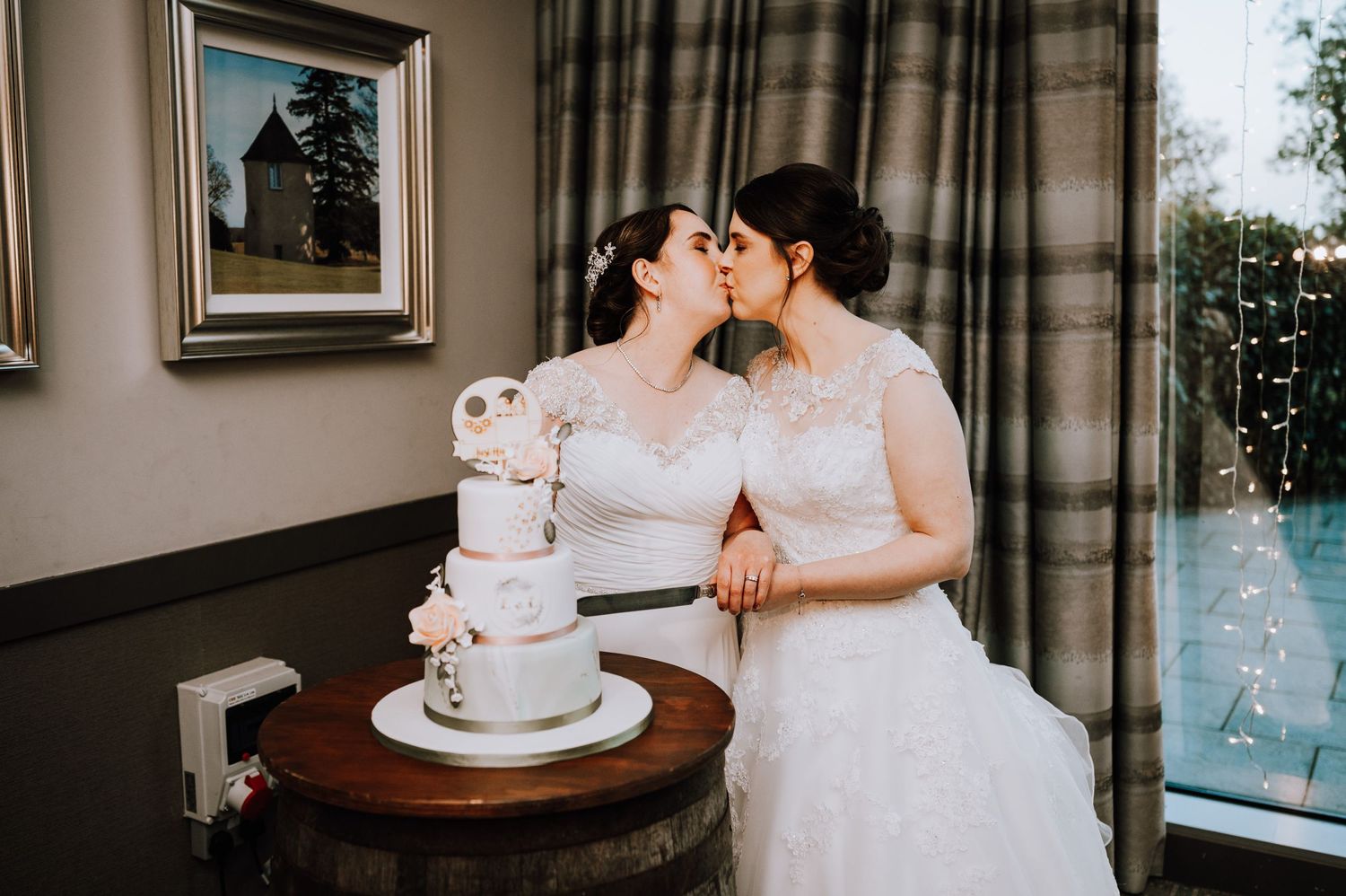 Two brides share an intimate moment next to their elegant white wedding cake in a beautifully decorated room.