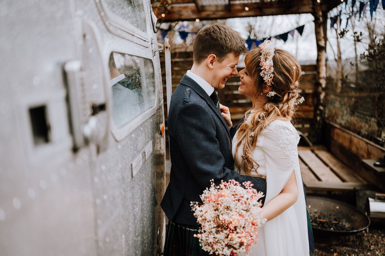 Intimate wedding portrait against vintage camper with bride holding pink bouquet and groom in traditional Scottish dress.