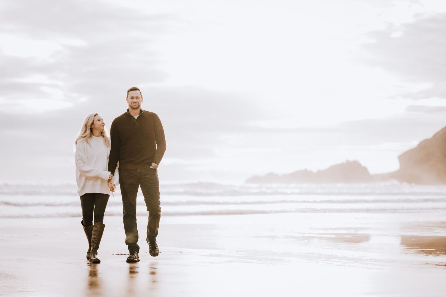 Couple walking together on wet sand with coastal cliffs in background.
