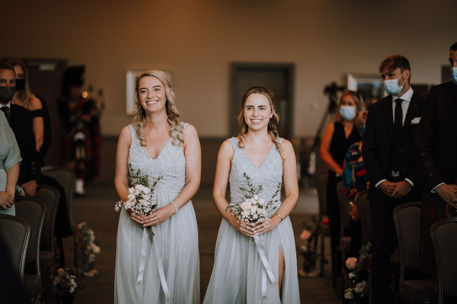 Bridesmaids in matching light grey gowns hold bouquets during indoor ceremony.