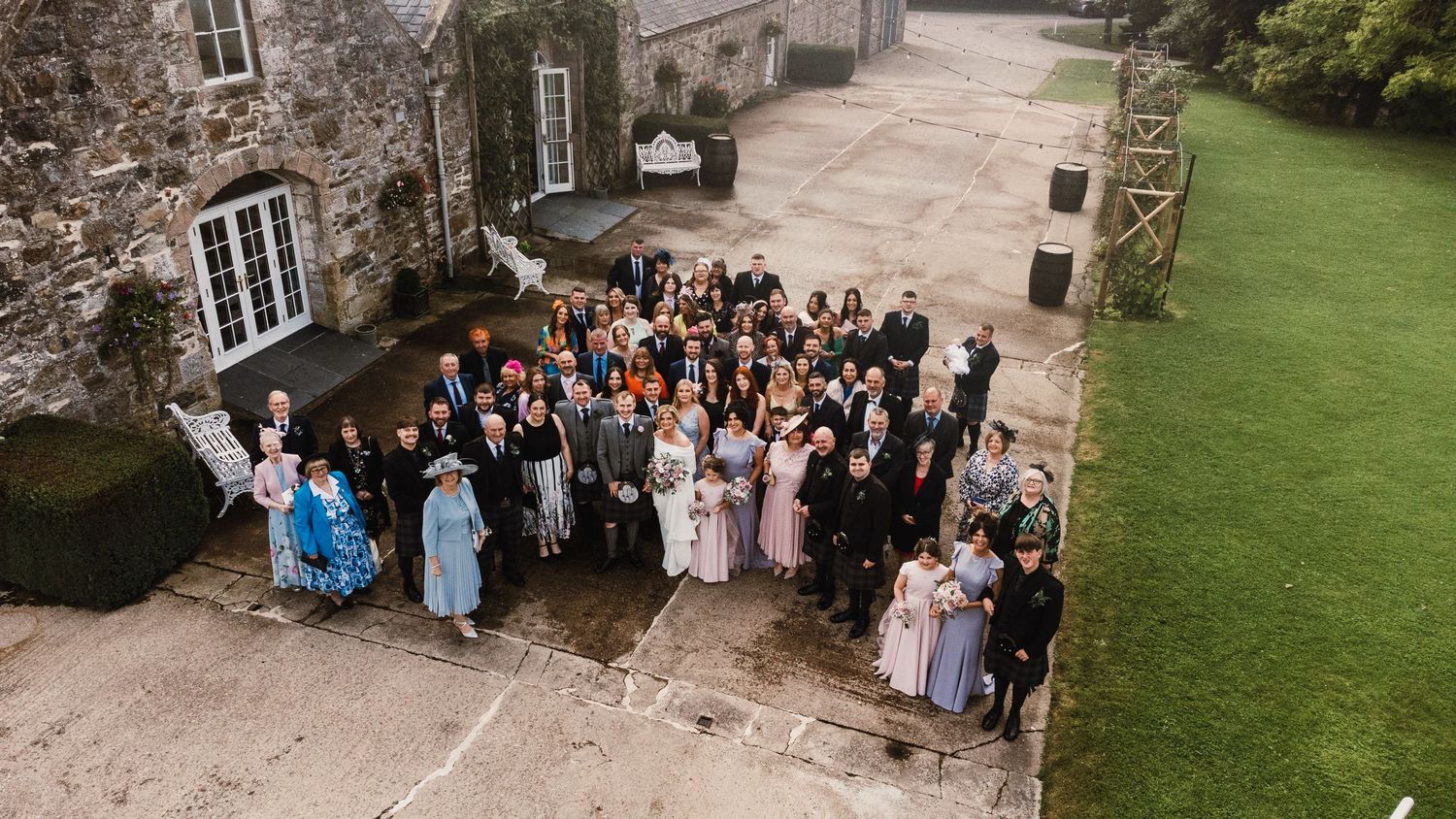 Aerial view of wedding guests gathered outside a stone building for a group photo.