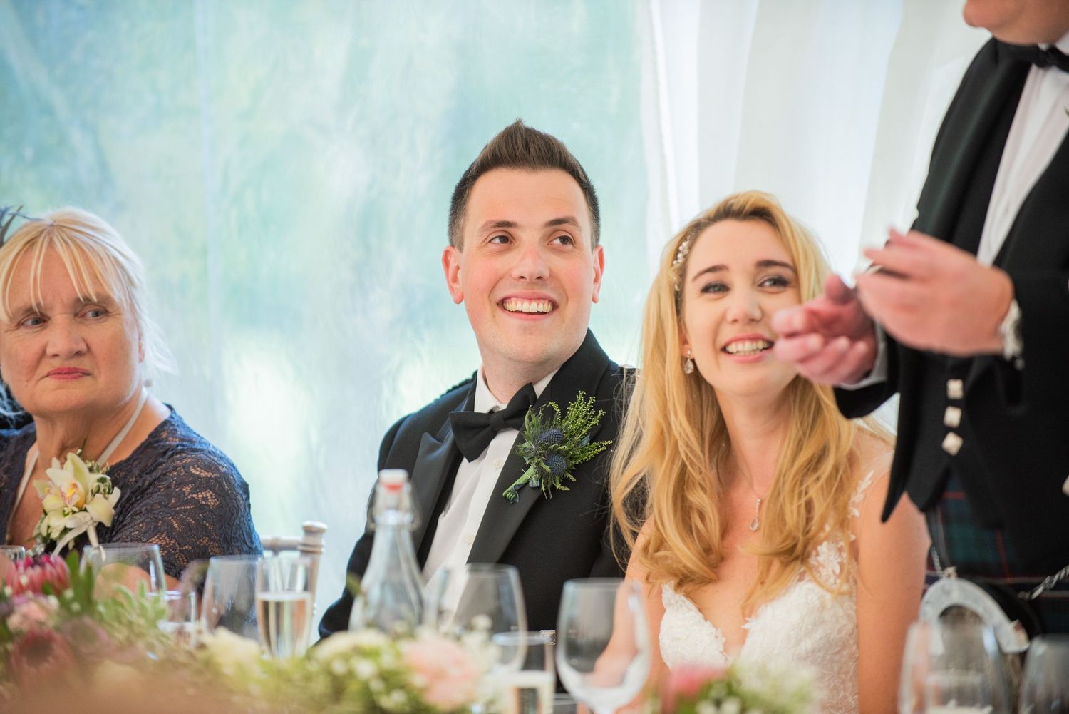 Newlyweds and guests share joyful laughter during wedding reception dinner in elegant white tent setting.