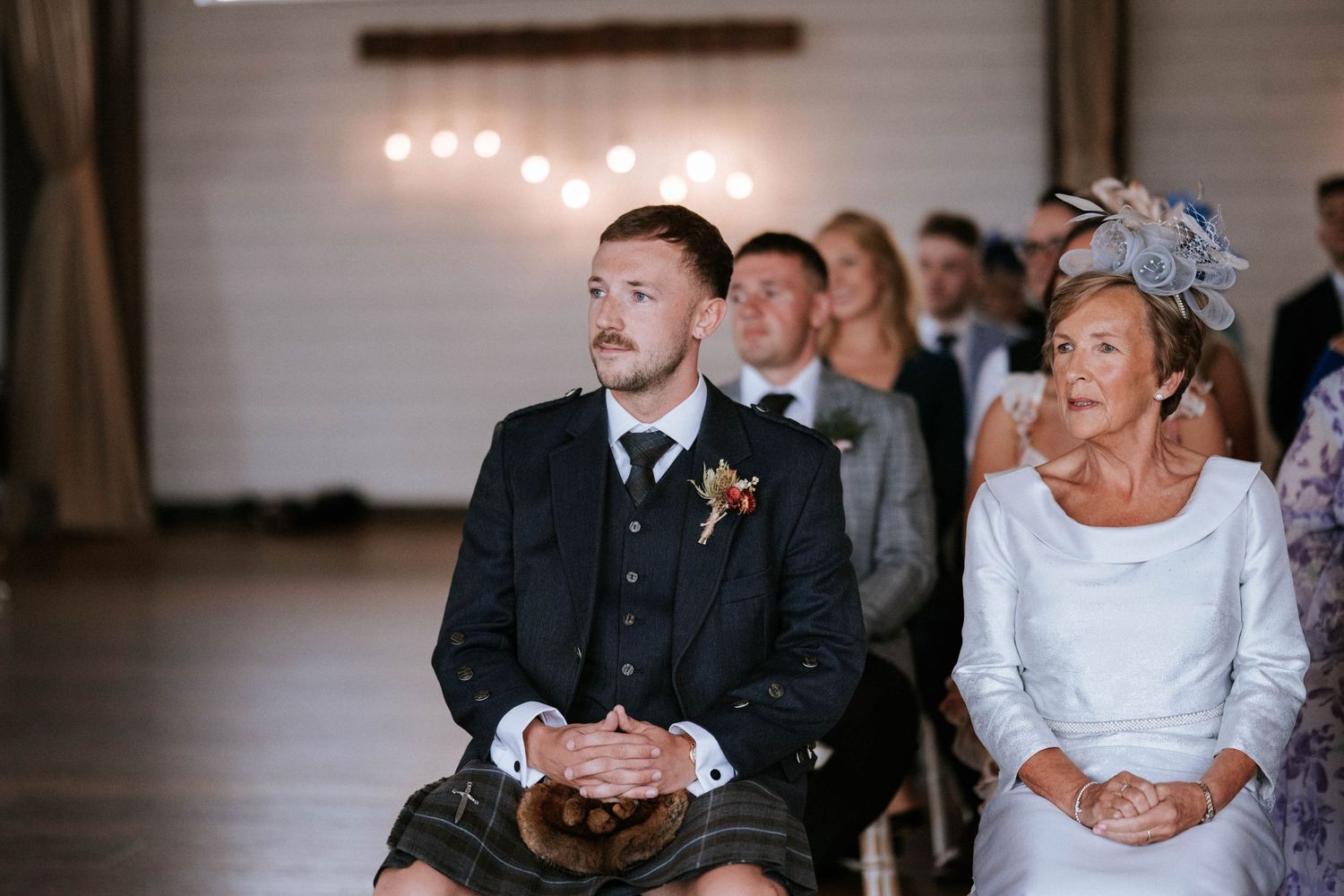 Wedding guests seated in rows watch attentively during an indoor ceremony with string lights overhead.
