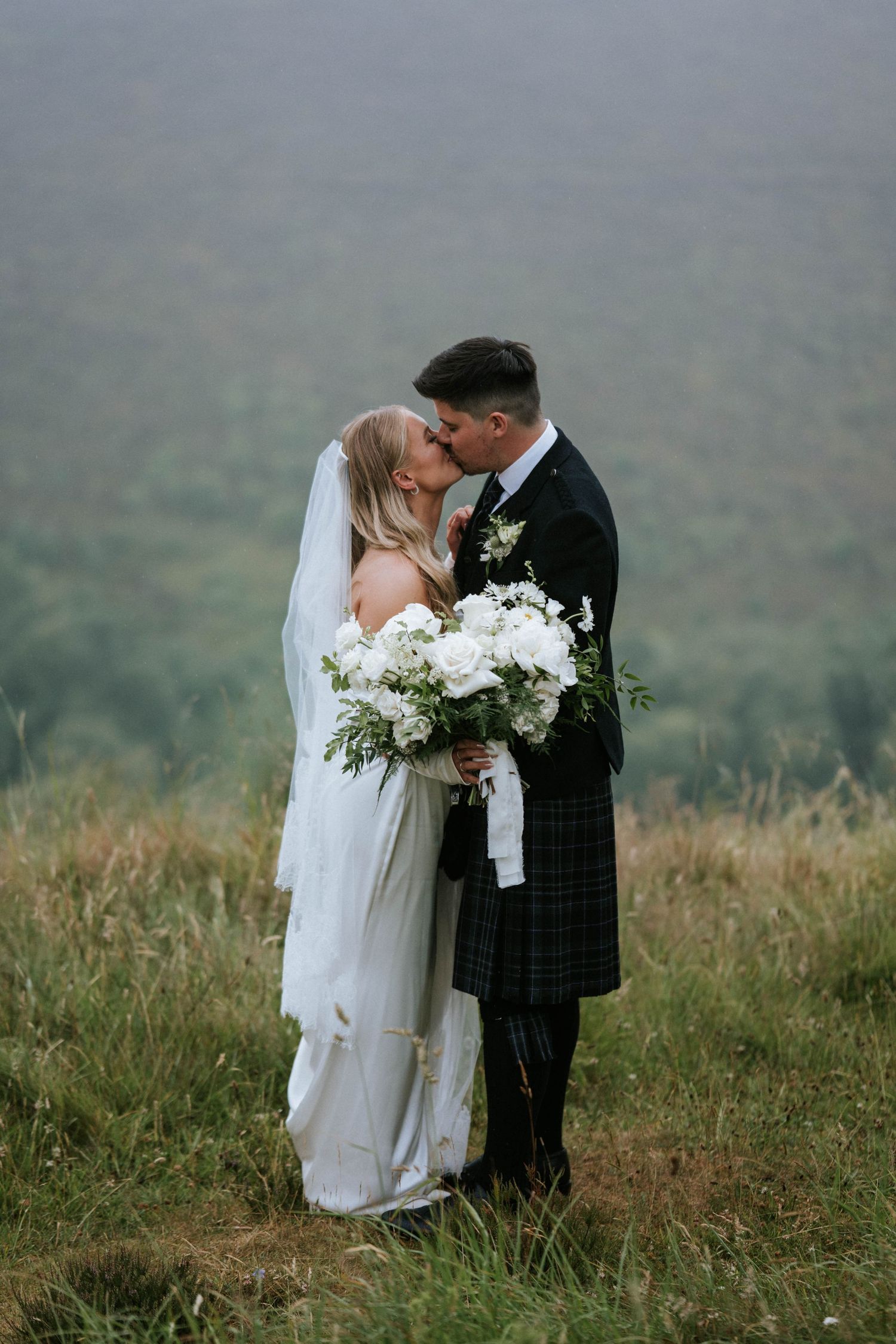 Newlyweds embrace in misty Scottish highlands with white floral bouquet.