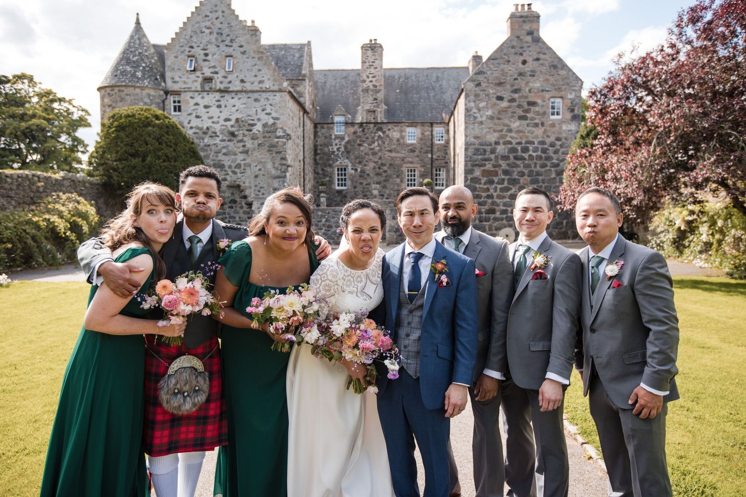 Wedding party poses together outside historic stone church building on sunny day.