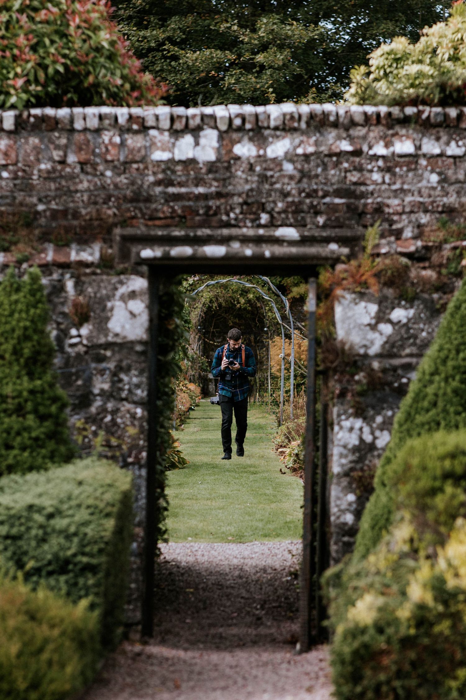 Stone archway frames a garden path through a rustic brick wall with autumn foliage in the background.