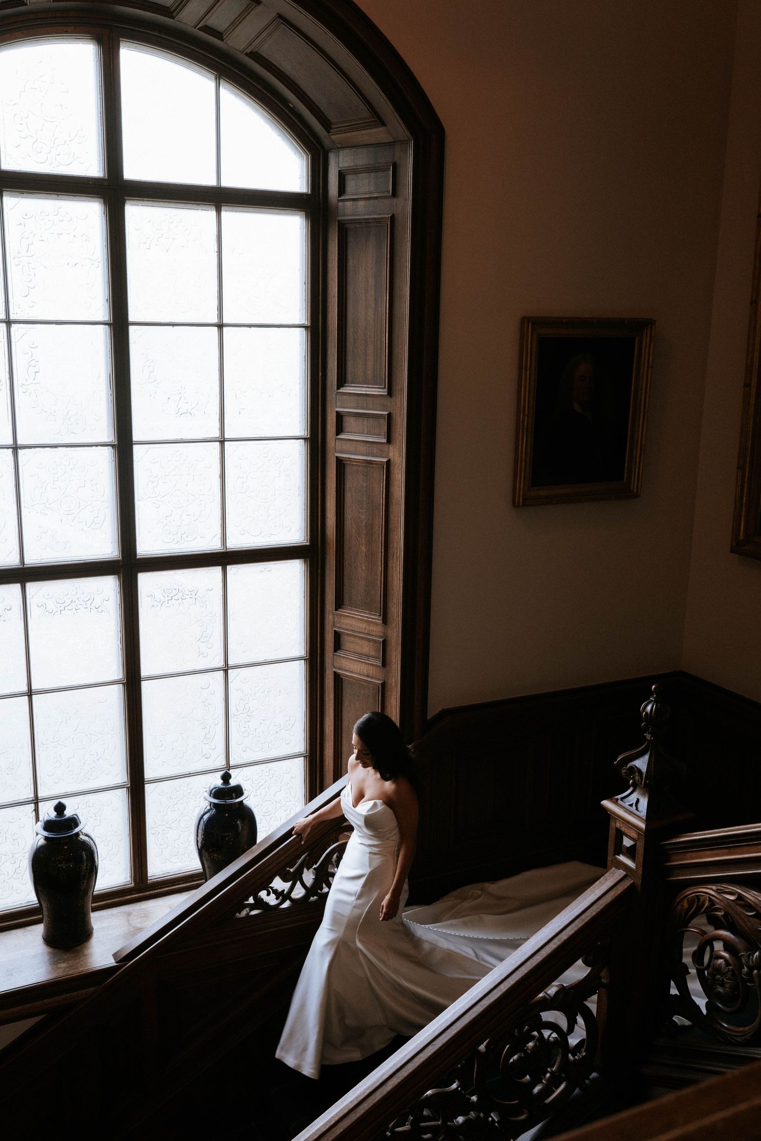 A bride in a white dress poses dramatically on dark wooden stairs by a large arched window.