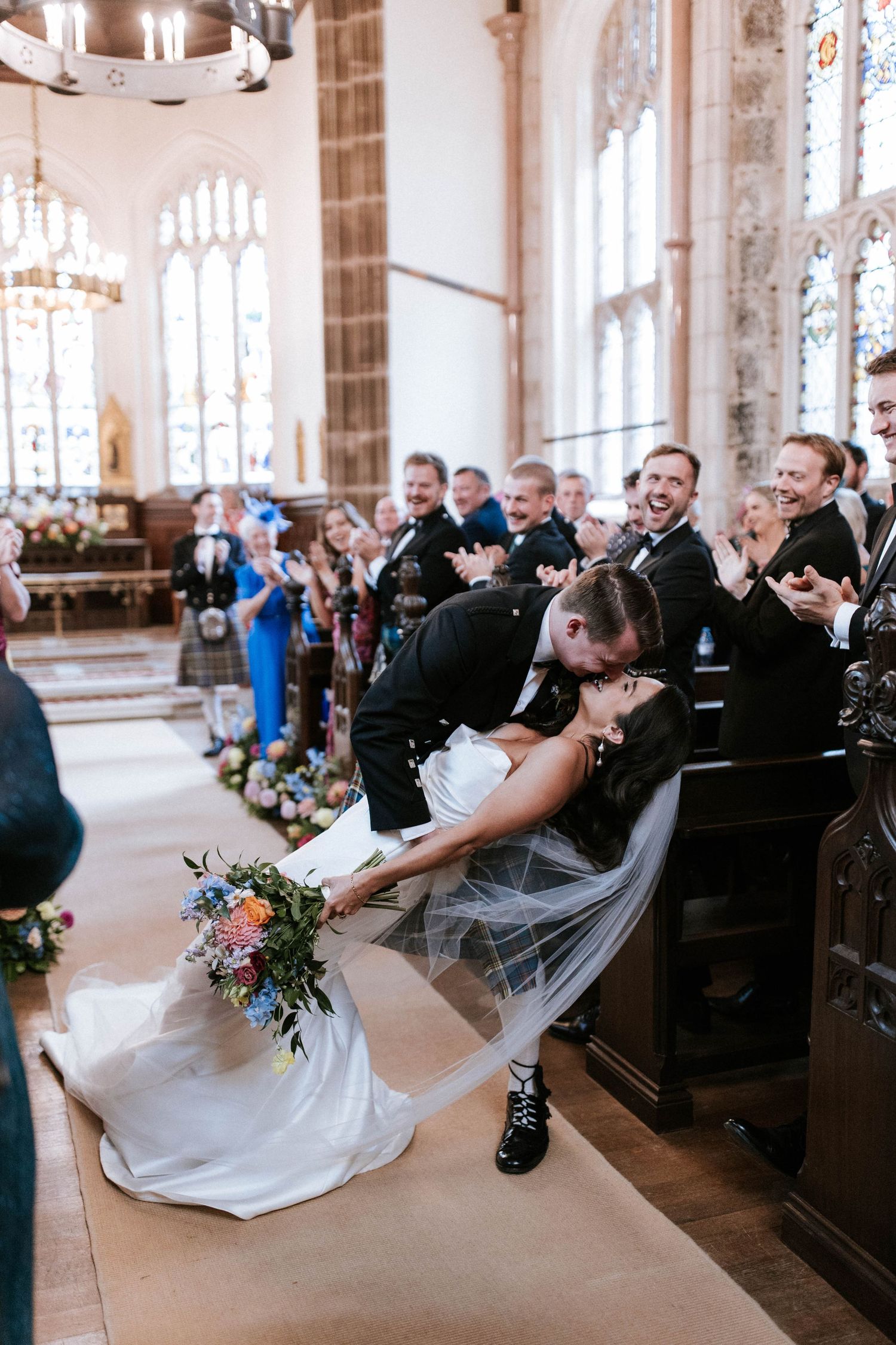 Romantic dip kiss during wedding ceremony in historic church with stained glass windows and wooden pews.