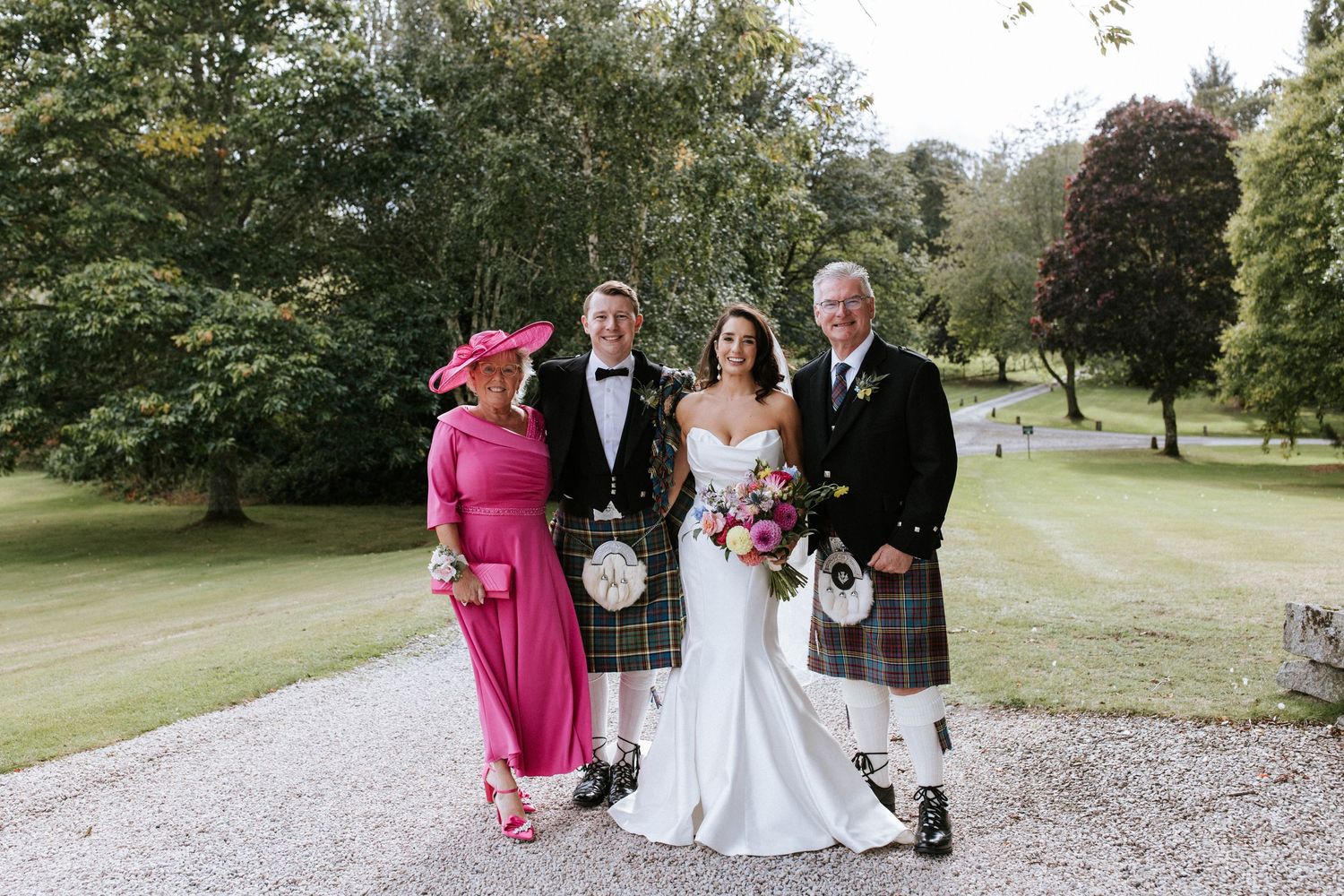 Intimate wedding group photo featuring Scottish formal wear and bright pink dress on garden grounds.