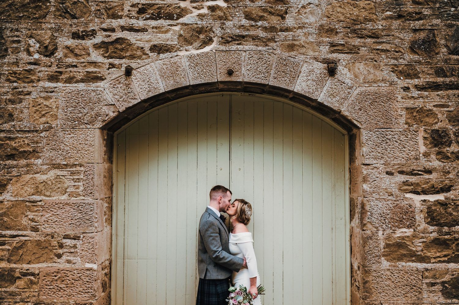 A couple shares an intimate moment against a cream-colored door framed by rustic stone walls at their Scottish wedding.