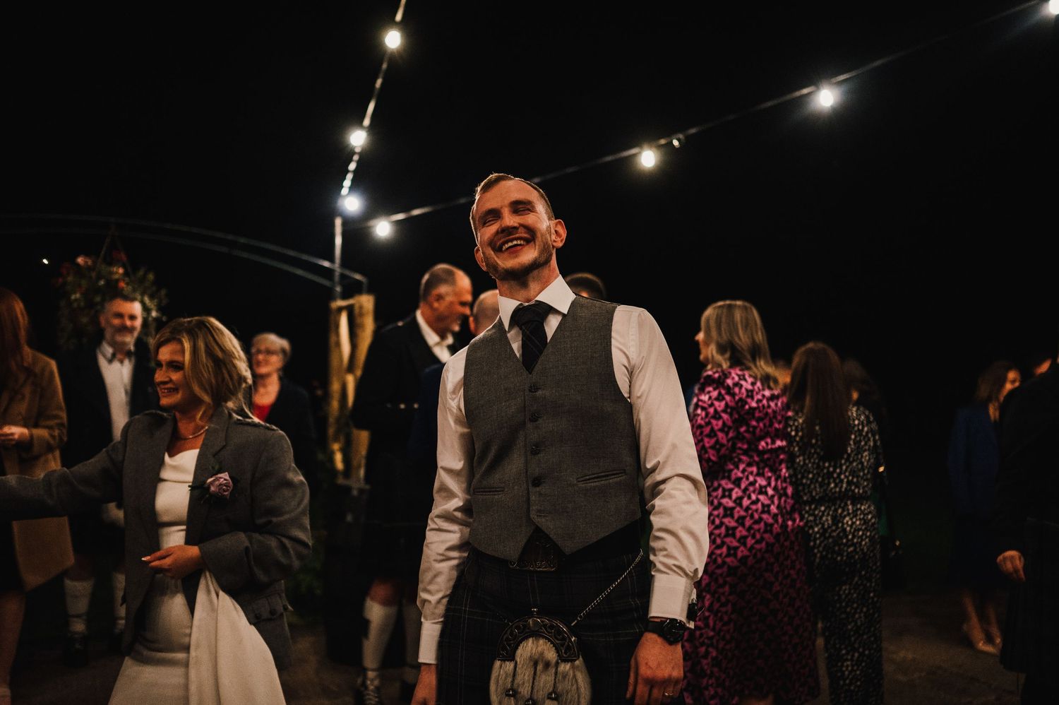Evening wedding reception under string lights with guests mingling and dancing.