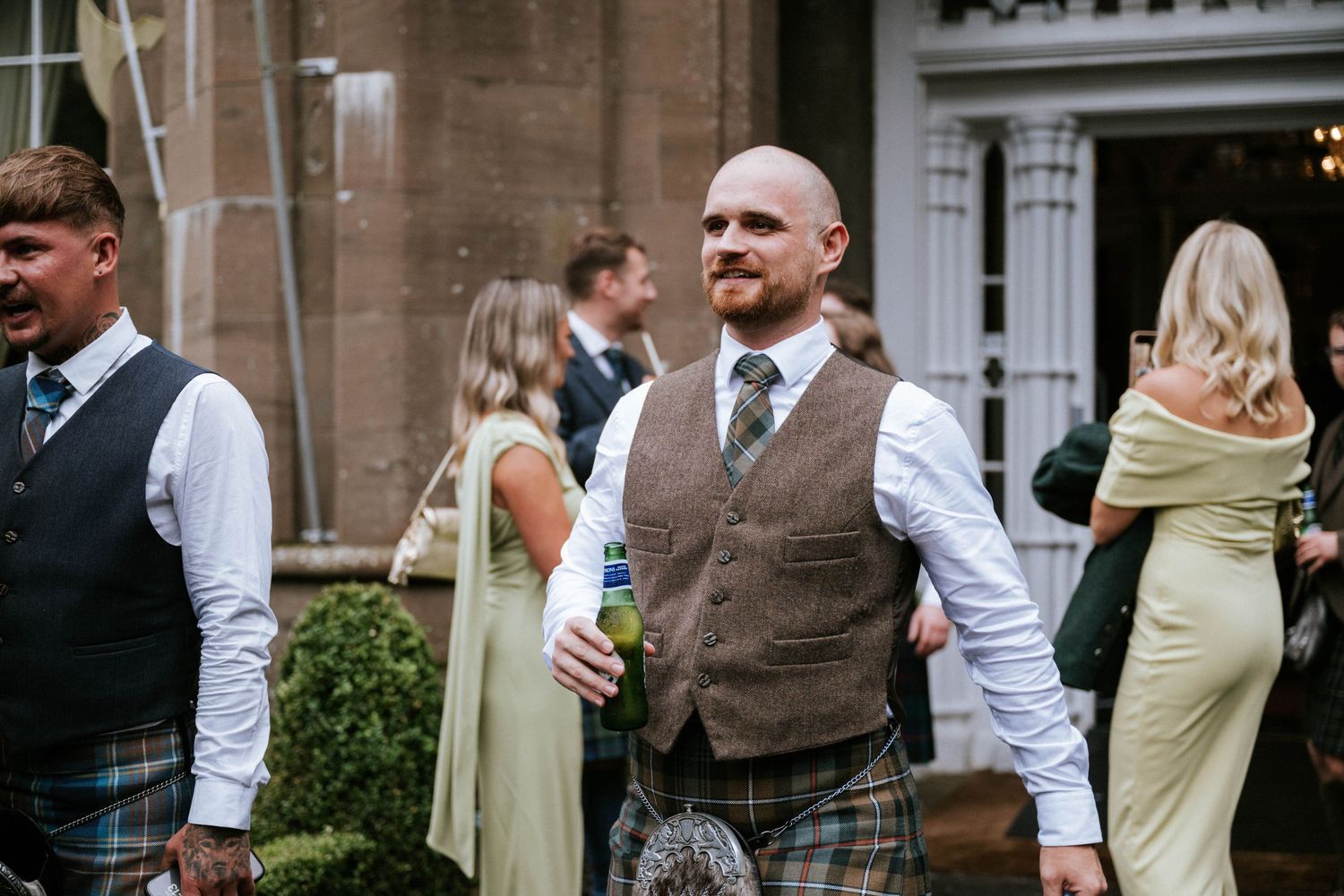 Wedding guests mingling outdoors wearing traditional Scottish kilts and formal attire.