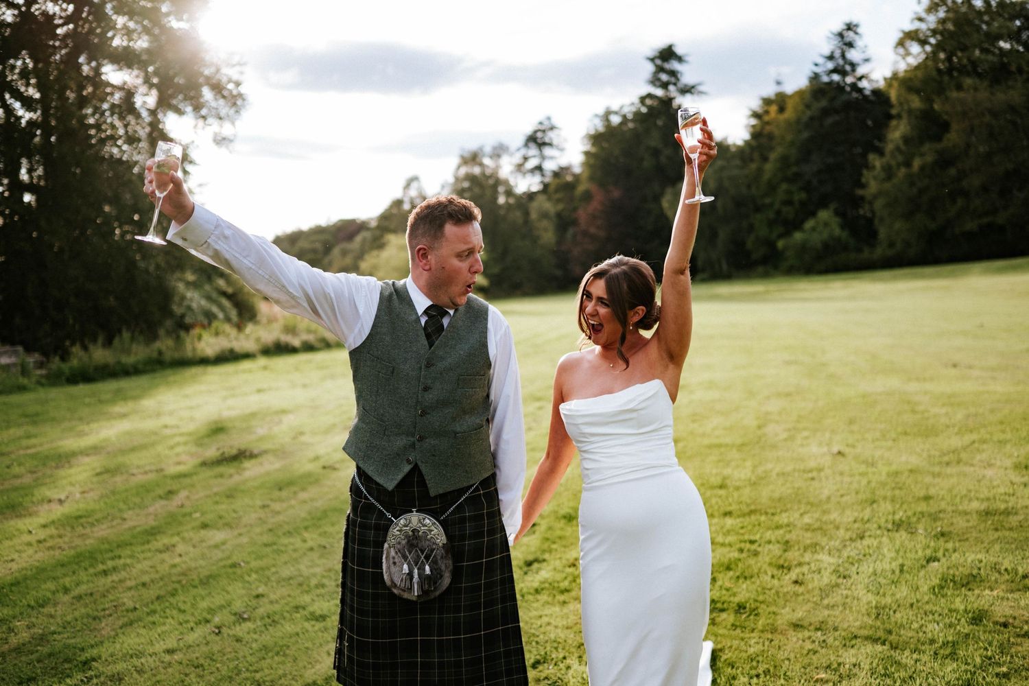 Aberdeen Wedding Photography. Newlyweds celebrate with raised arms on a sunlit grassy hill, groom wearing traditional Scottish kilt.