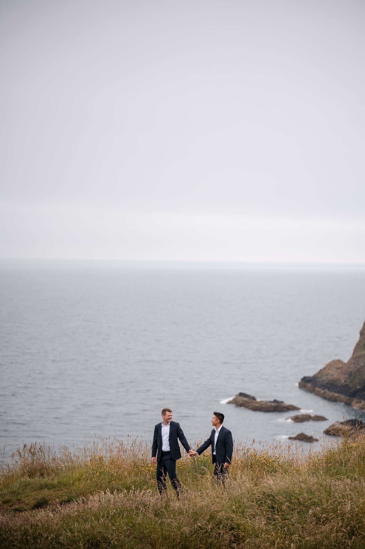 Same Sex Wedding Photography. Two figures stand together on a grassy cliff against a moody seascape with rocky outcrops.