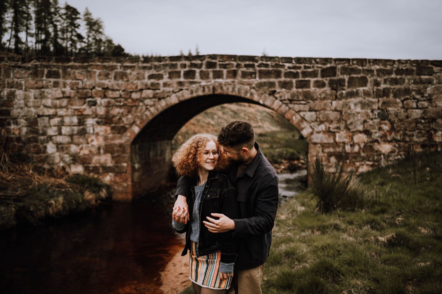 Engagement photography session Aberdeen. Two people embrace near an ancient stone wall with pine trees in the background.