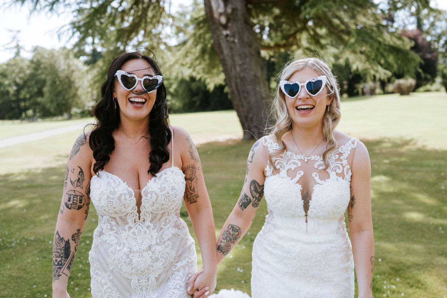 Aberdeen Wedding Photography. Two brides in white dresses and sunglasses smile together on a sunny lawn.