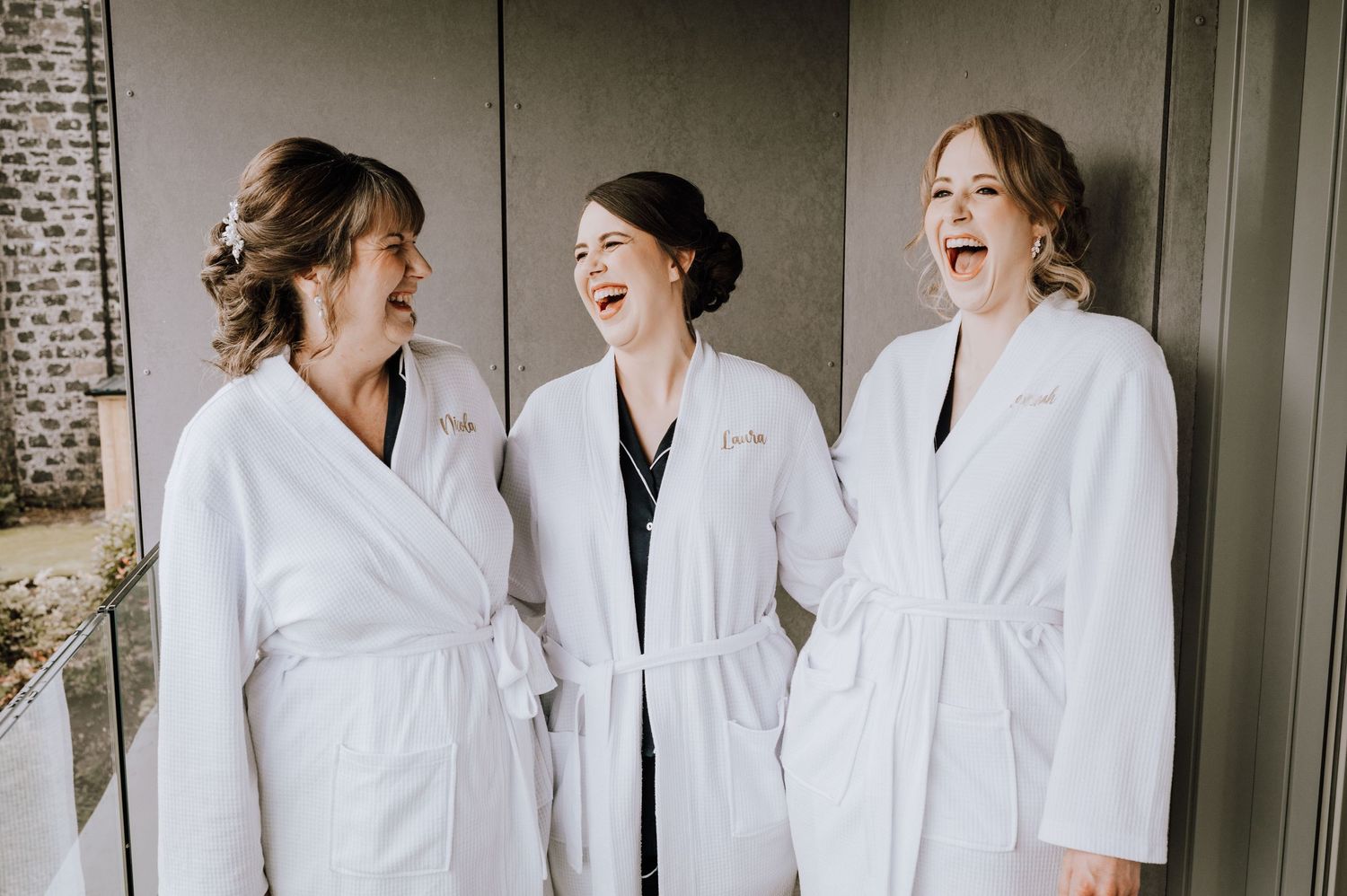 Bridal party in white robes sharing laughs against brick wall. Meldrum House Wedding Photography.