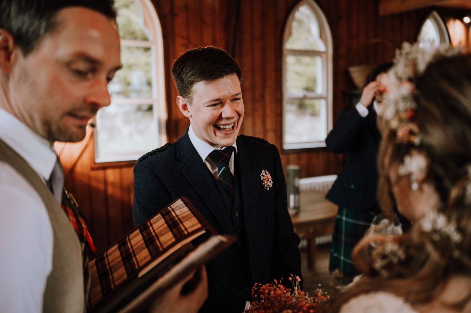 A candid moment during a wedding ceremony with guests laughing in a wood-paneled room.