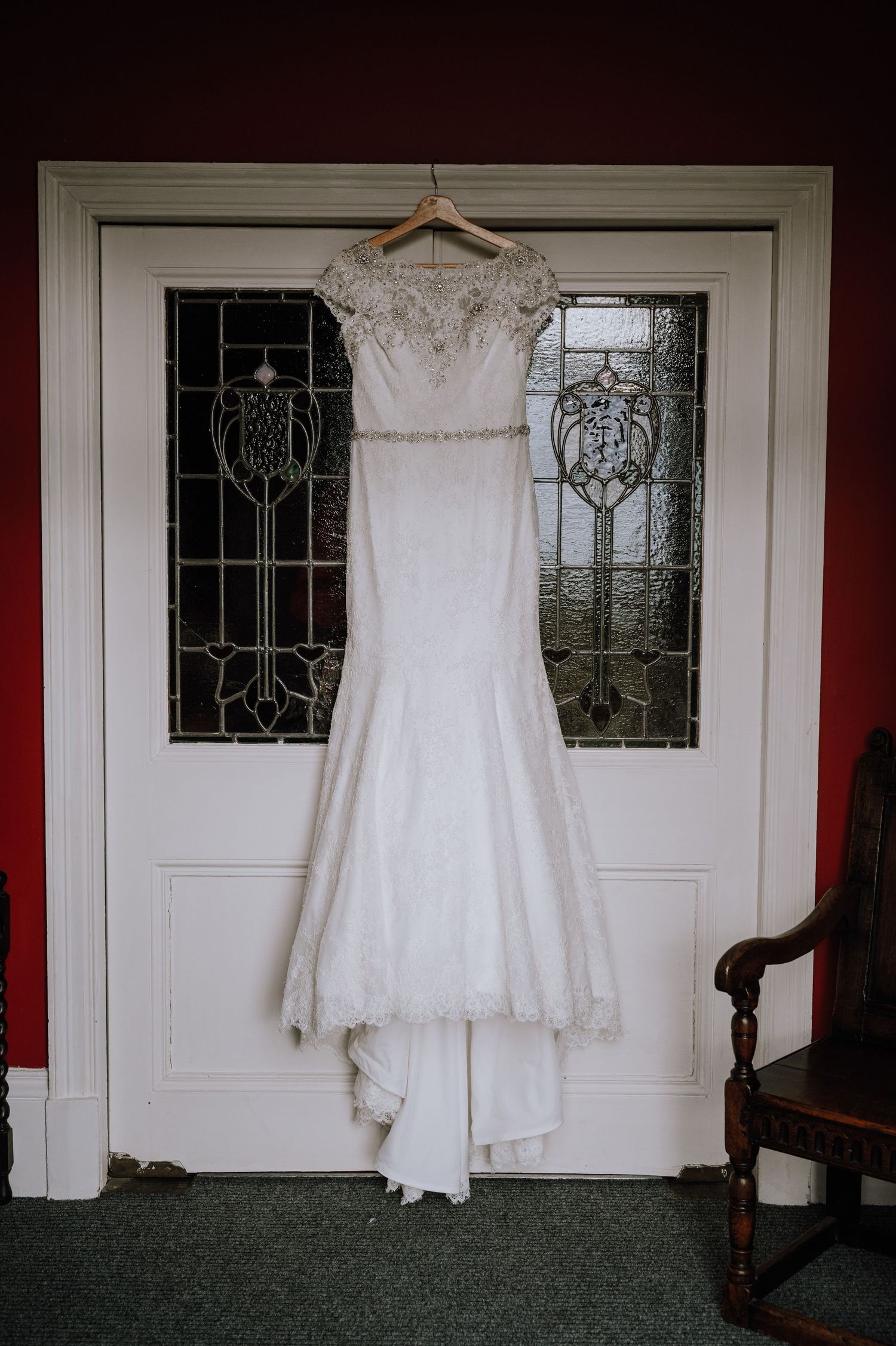 An elegant white lace wedding dress hangs against stained glass doors in a traditional setting.