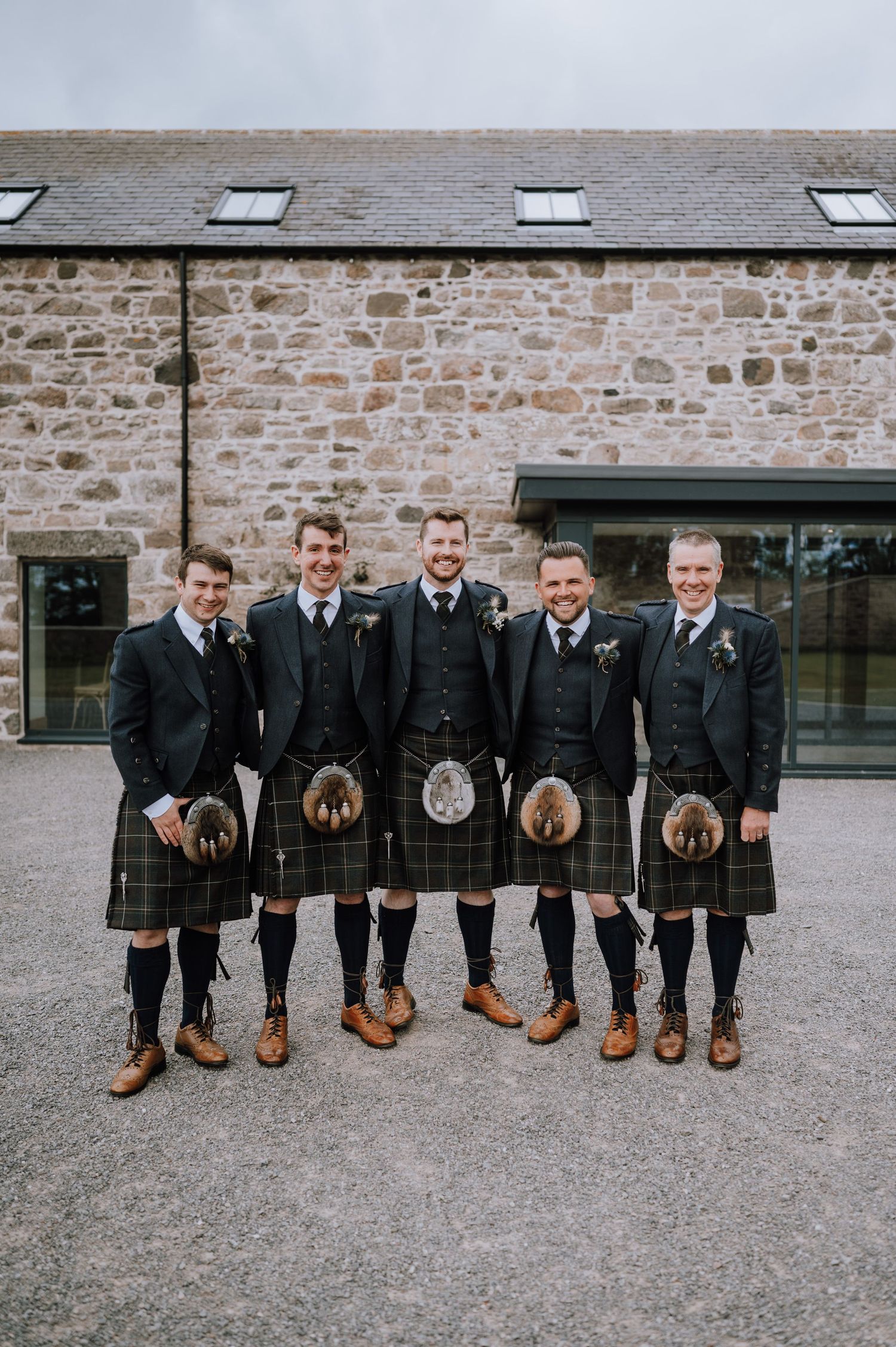 Five groomsmen in matching kilts and formal wear stand in front of a rustic stone building.