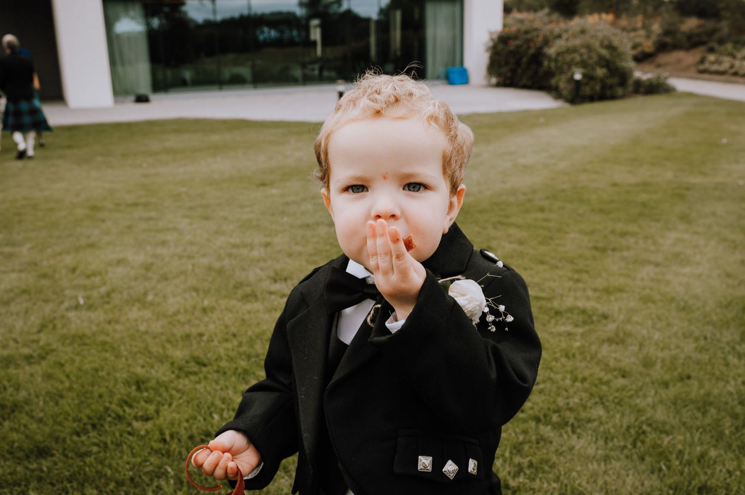 Young child in formal black attire with white boutonniere at outdoor wedding event.
