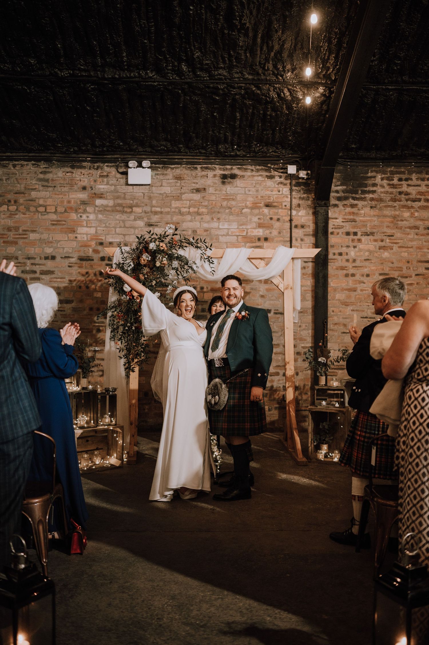 A wedding celebration in a rustic brick venue with glowing lights and Scottish kilts.