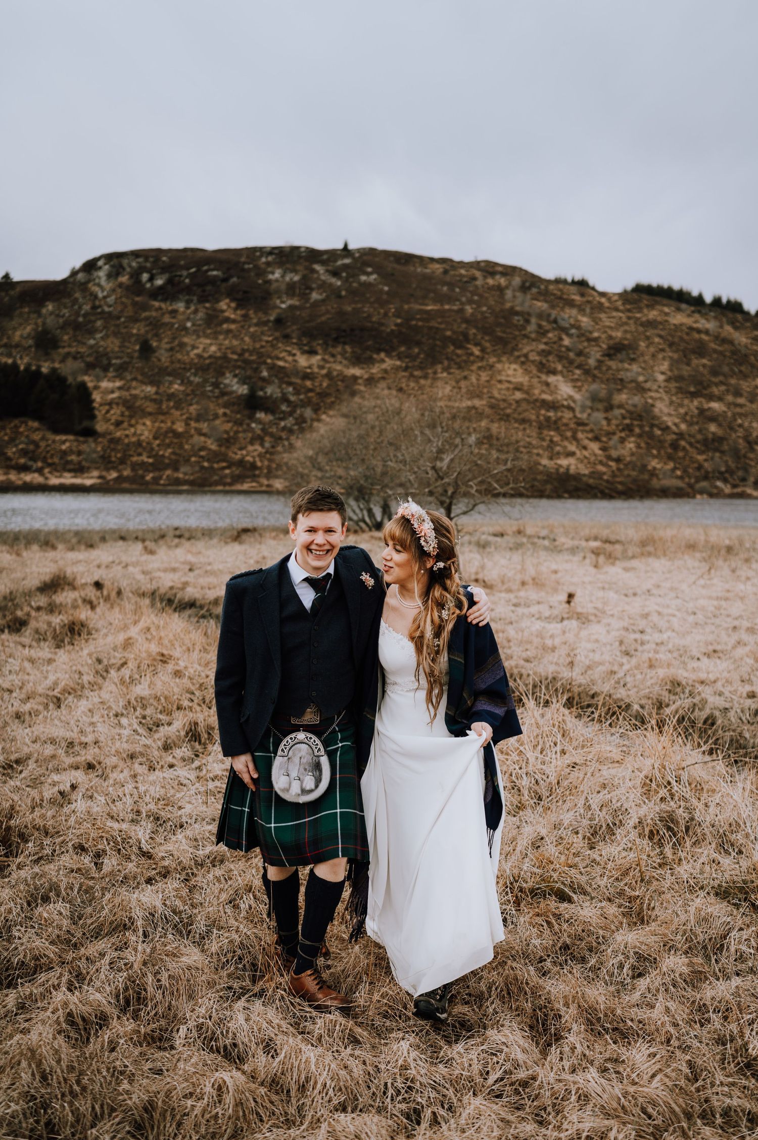 Couple in wedding attire stands in Scottish highlands with mountains and lake in background.