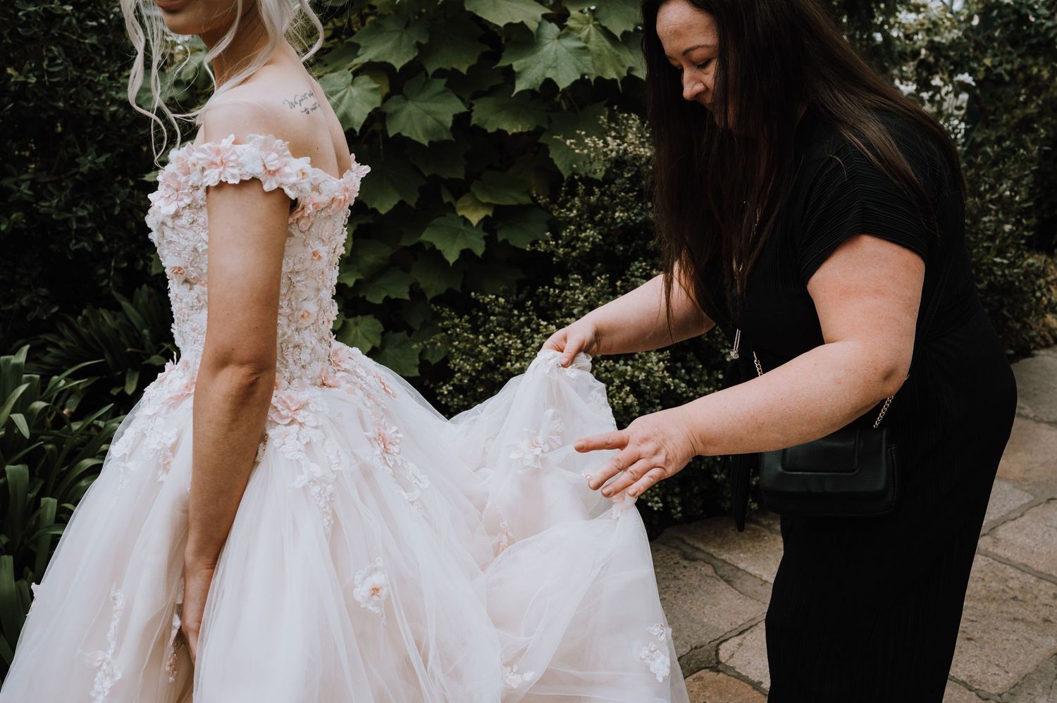 A delicate white wedding gown with floral details being adjusted in a garden setting.