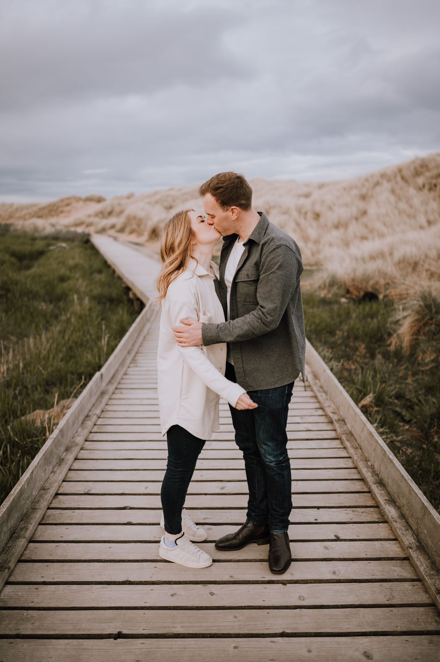 A couple shares a romantic moment on a wooden boardwalk surrounded by coastal dunes.