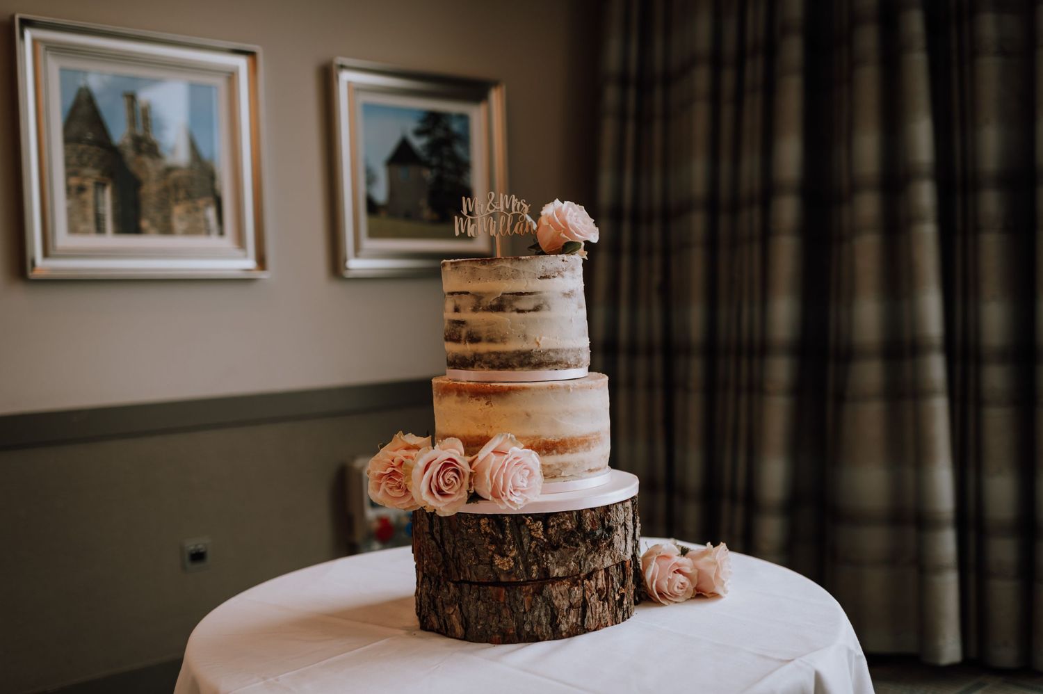 Semi-naked wedding cake decorated with pink roses sits on a rustic wood slice stand.