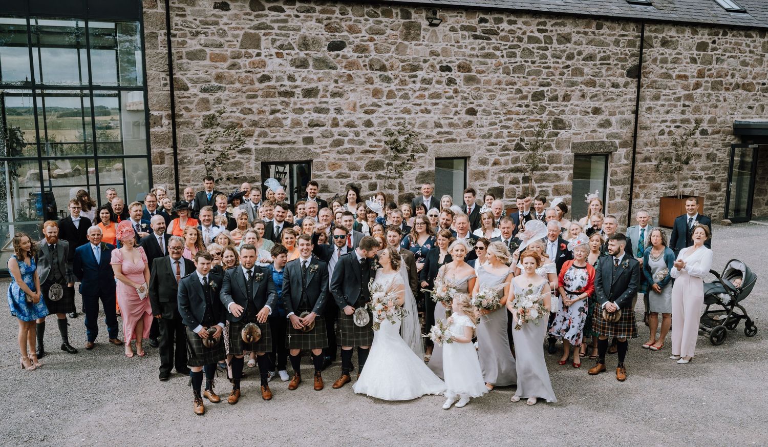 Large wedding party group photo in front of rustic stone building during outdoor celebration.