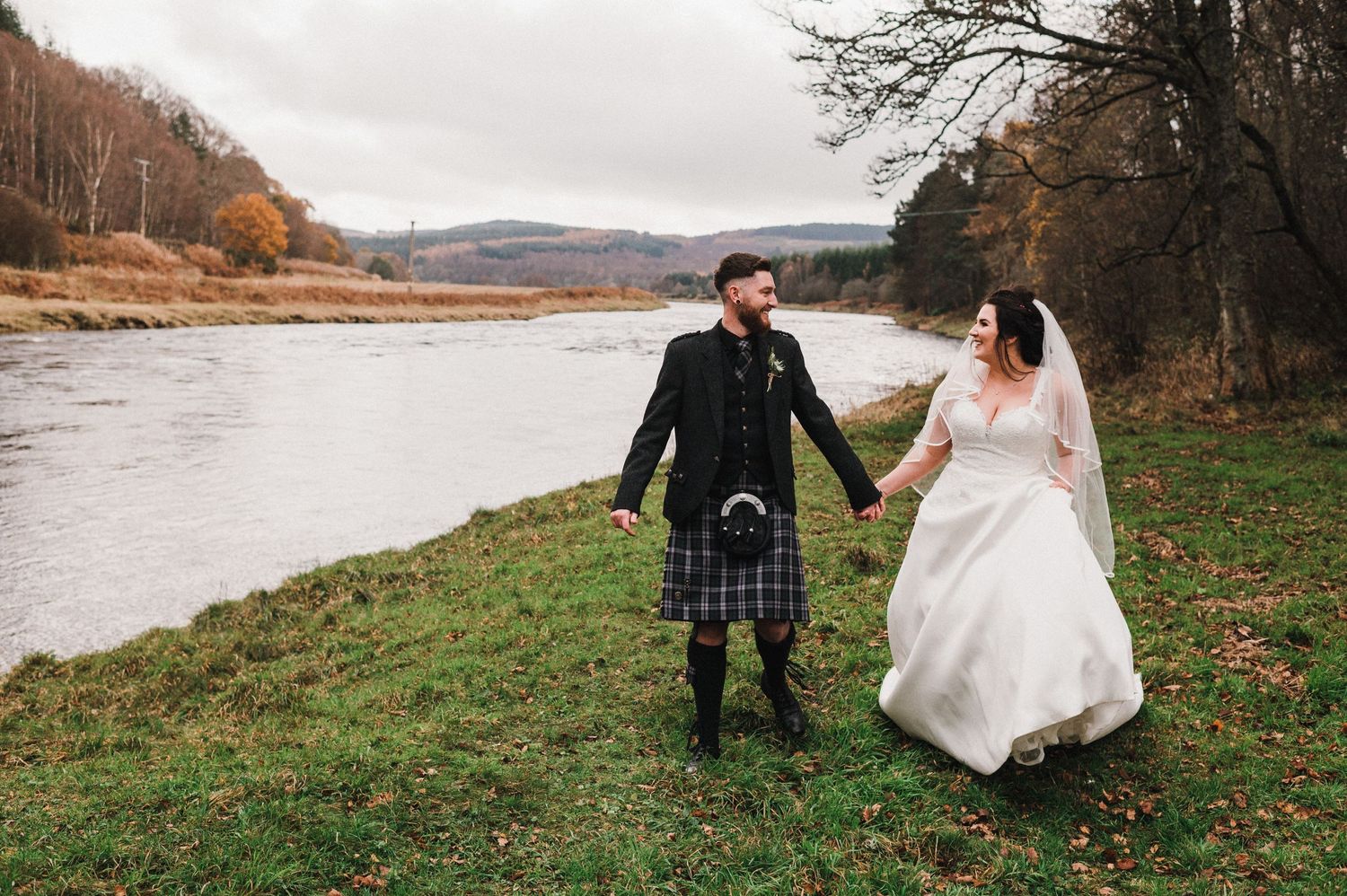 Wedding couple walks along riverside, groom wearing traditional Scottish kilt.