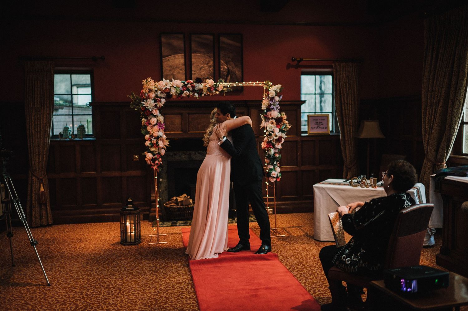 A wedding ceremony silhouetted against windows under a flower-adorned arch with red carpet.