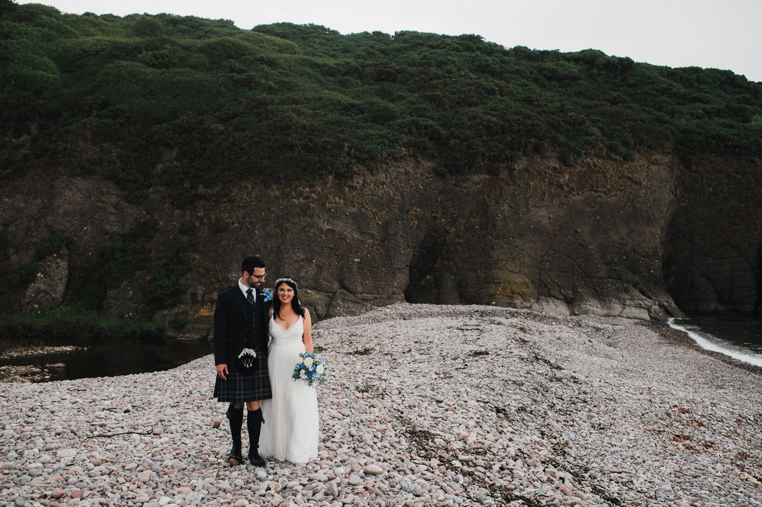 A coastal wedding portrait with cliffs and pebble beach creating a dramatic backdrop.