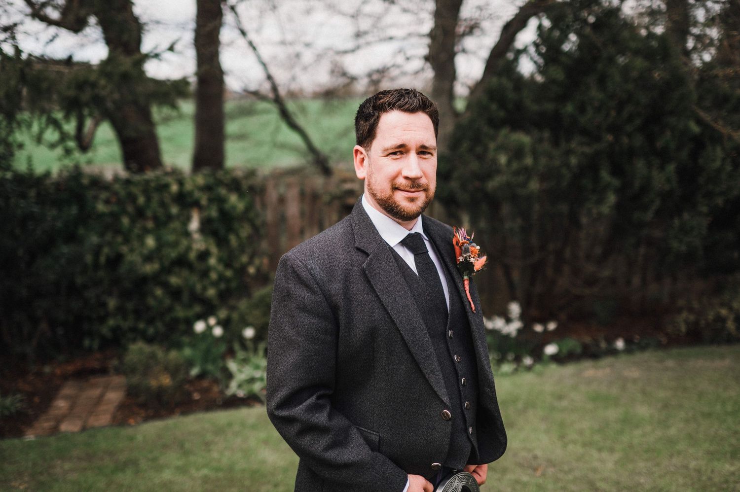 Groom in traditional Scottish attire with kilt and formal jacket poses in a garden setting.