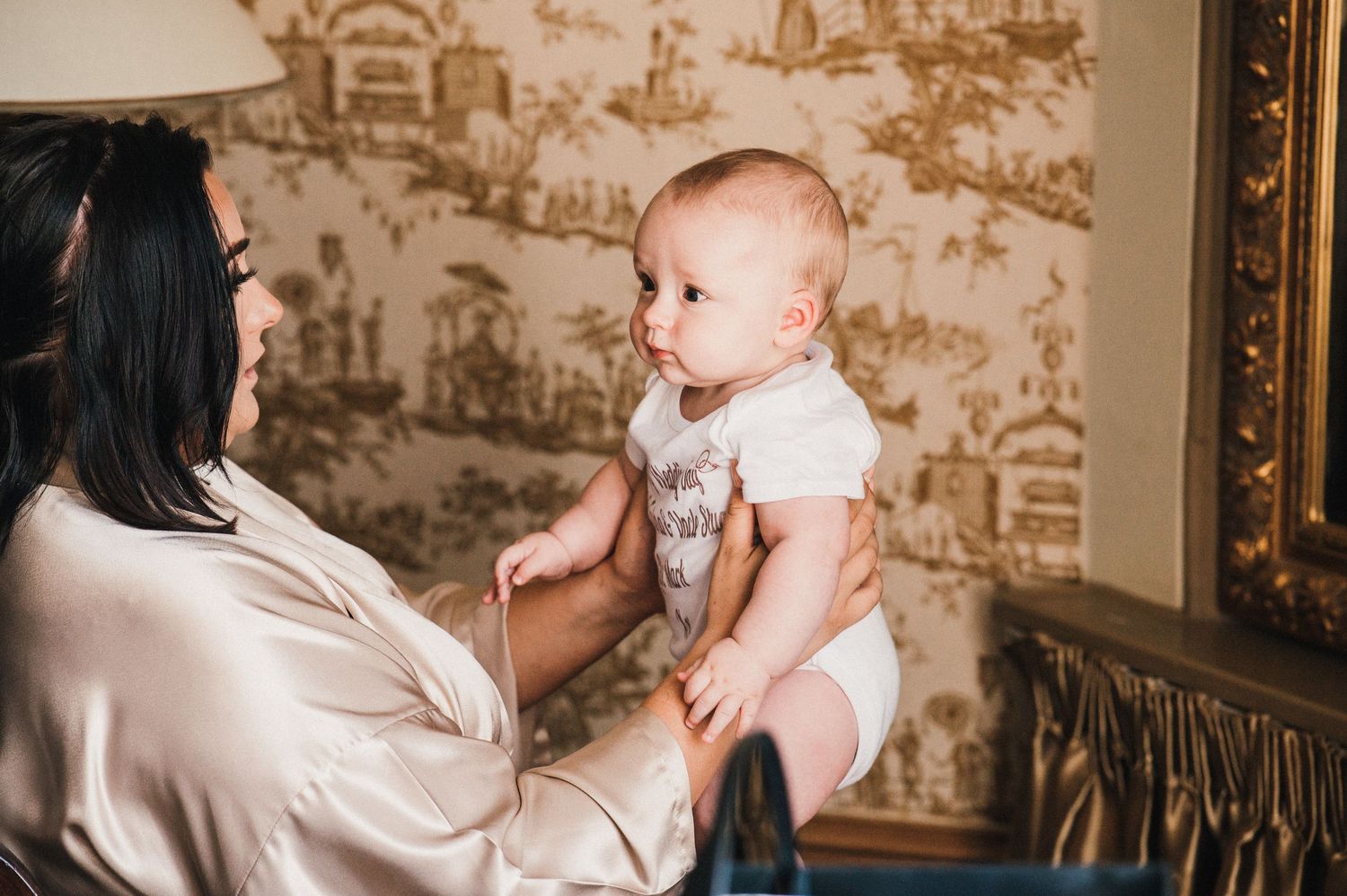 Adult holding a baby in front of decorative wallpaper in an elegant room.