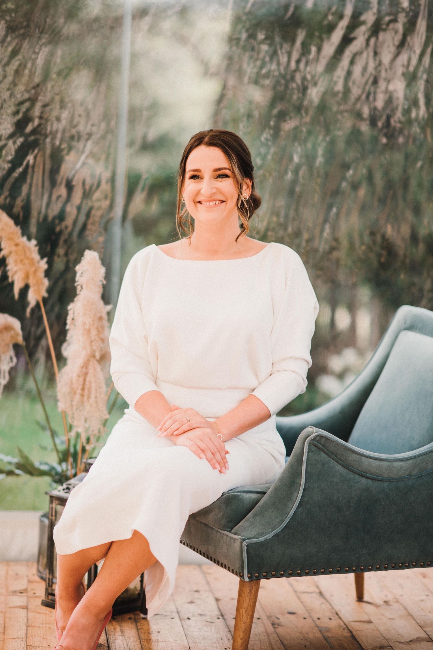 Elegant bride in white dress sits on modern gray velvet chair with pampas grass decor.