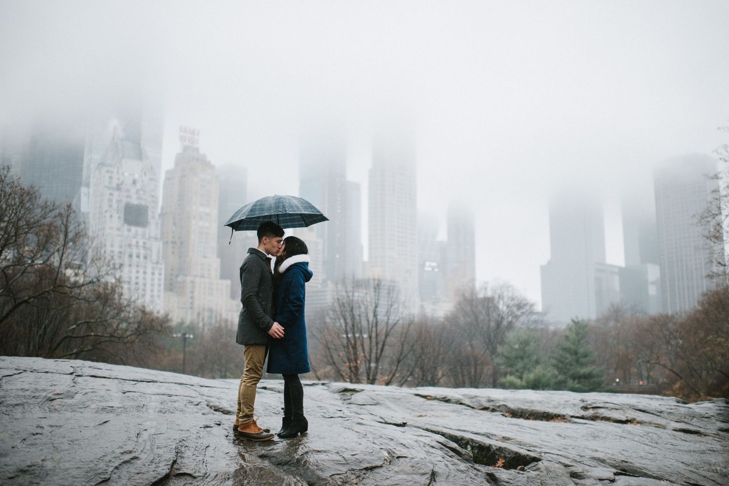 A romantic couple shares an umbrella in snowy Central Park with foggy Manhattan skyscrapers in the background.
