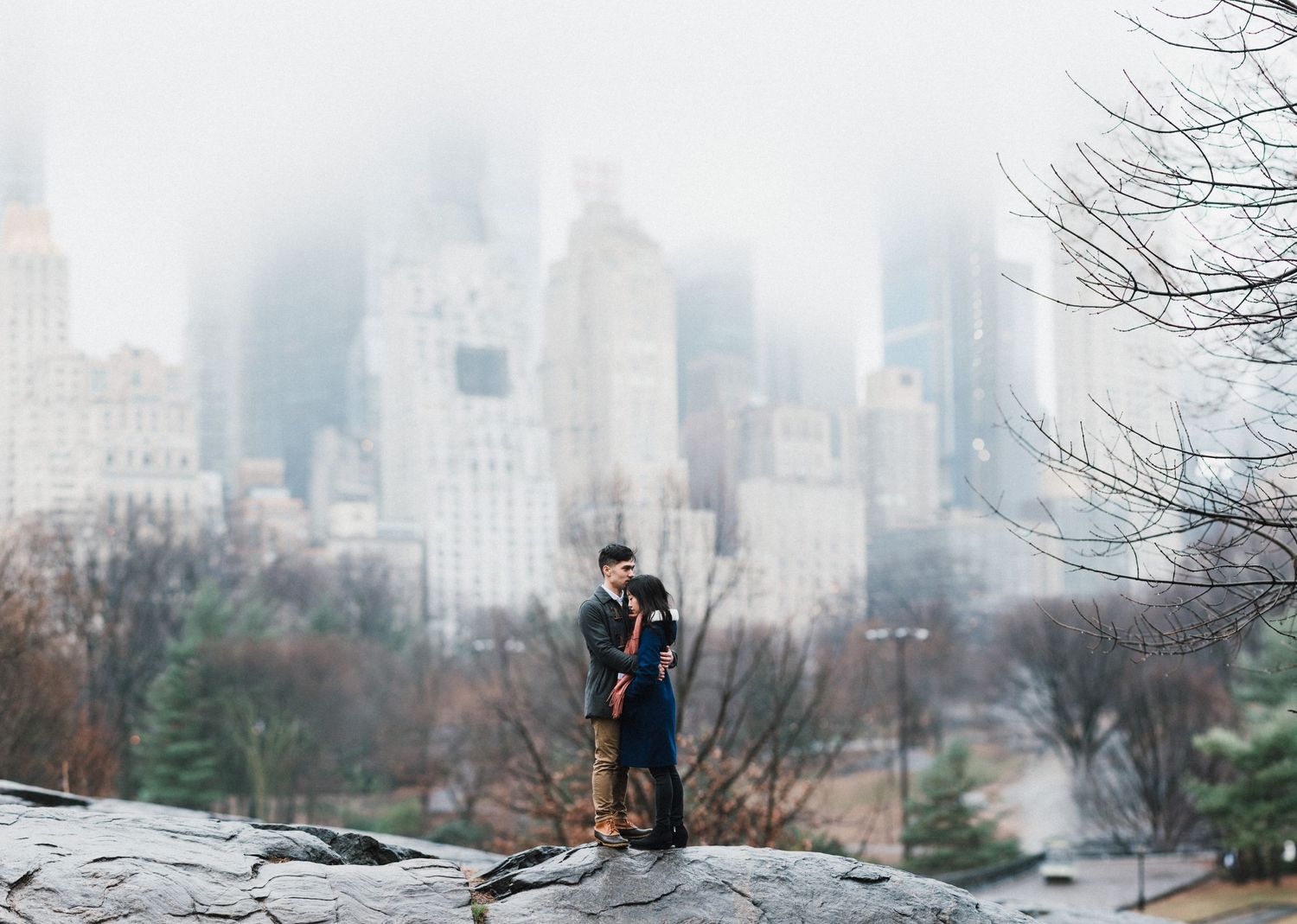 A couple stands on rocky terrain with misty Manhattan skyscrapers in the background.